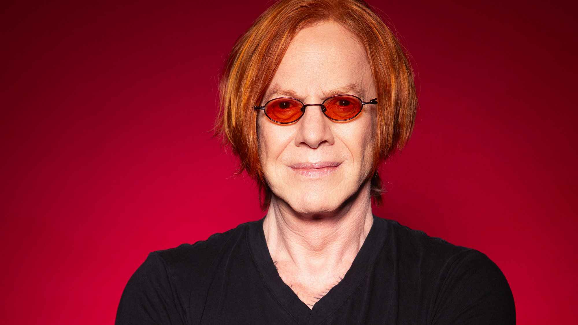 A man with red hair and black v-neck t-shirt against a deep red background wearing red sunglasses