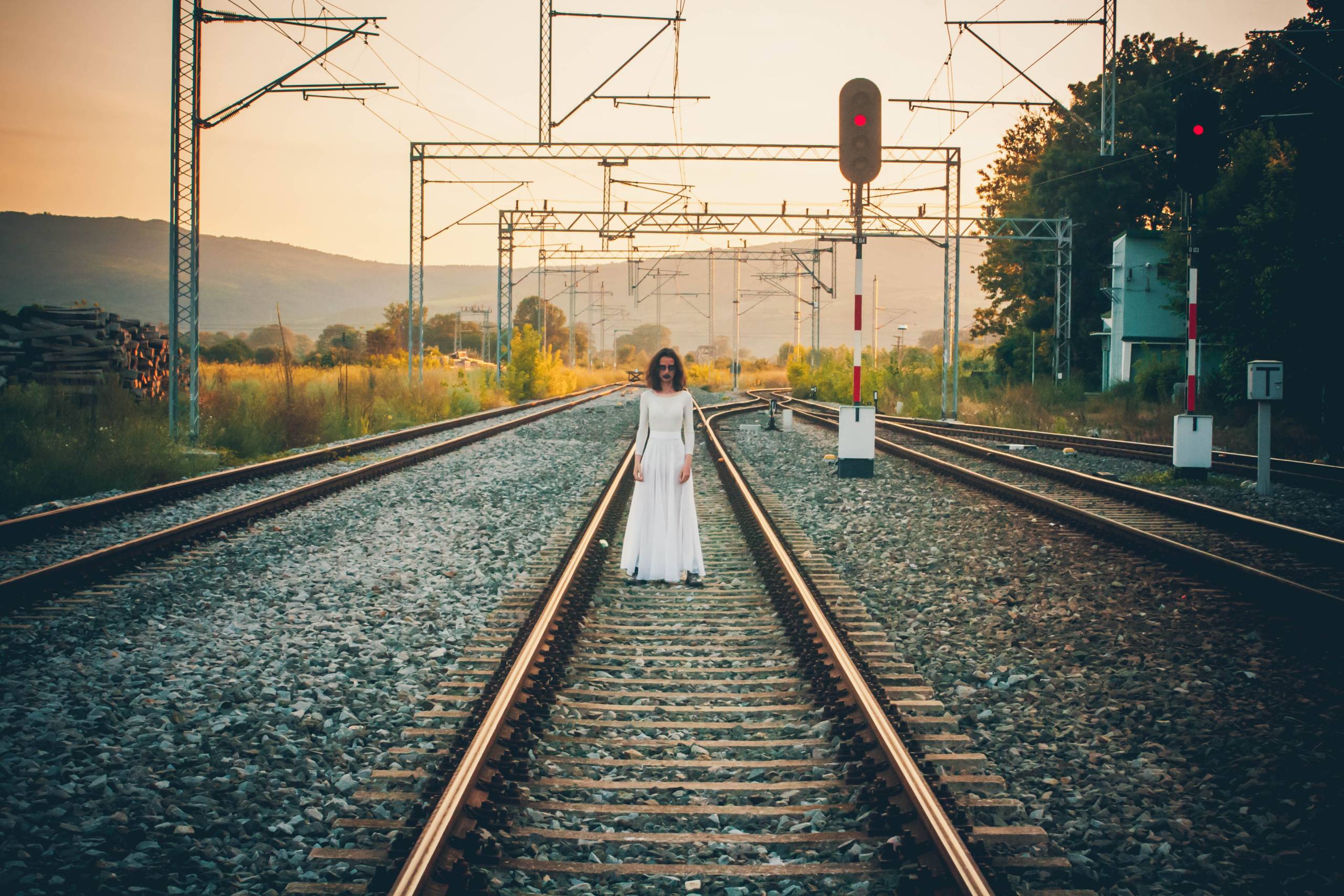 A ghostly woman wearing a long white dress stands on some train tracks, at a distance.