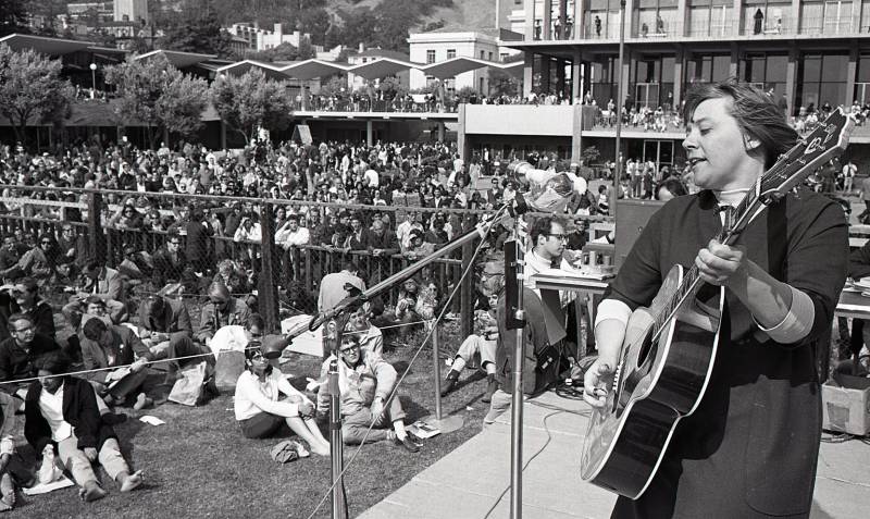 woman holding guitar sings into mic in front of large crowd