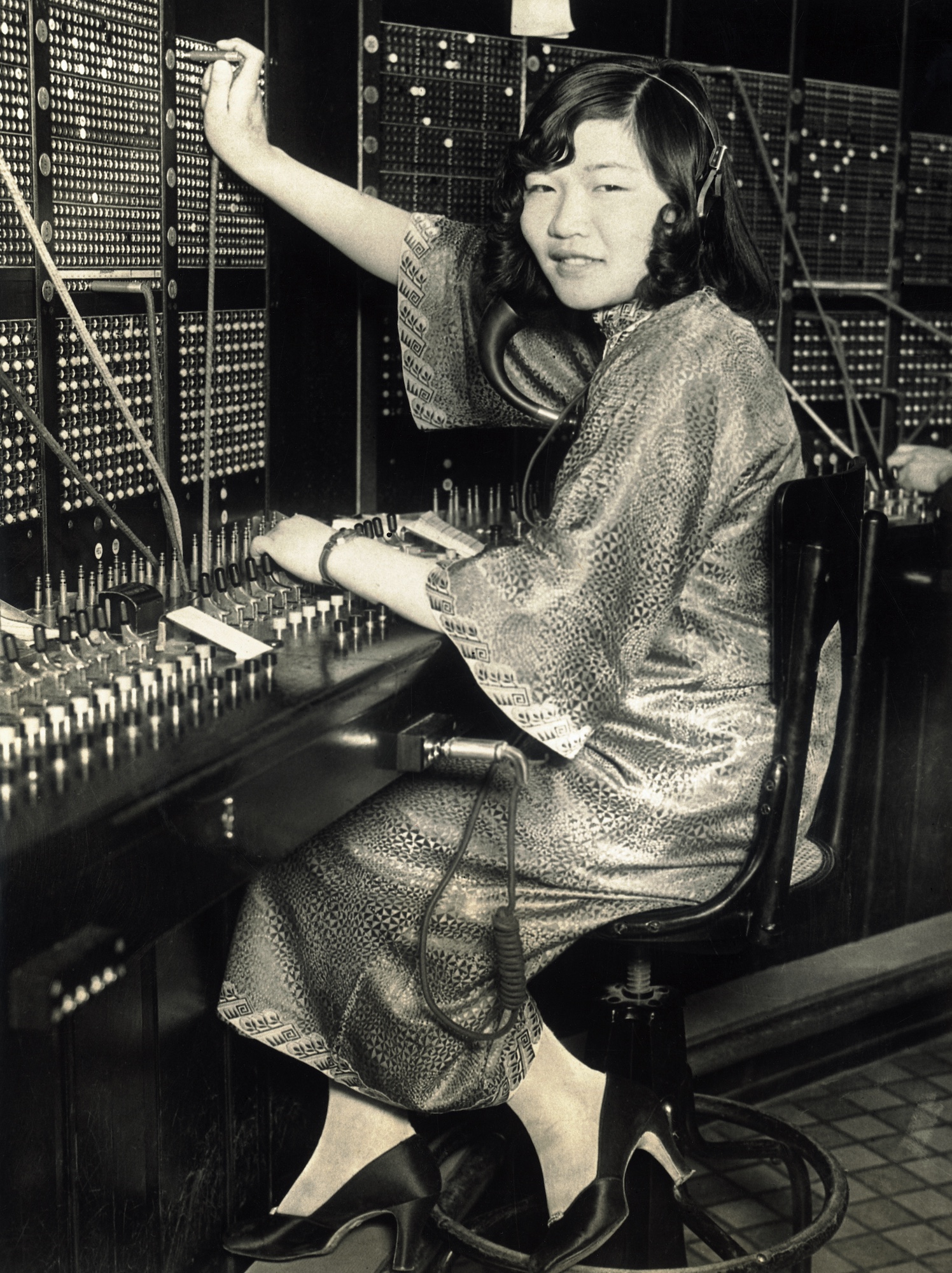 A young Asian woman operates a switchboard, while wearing a silk gown and telephone headset.