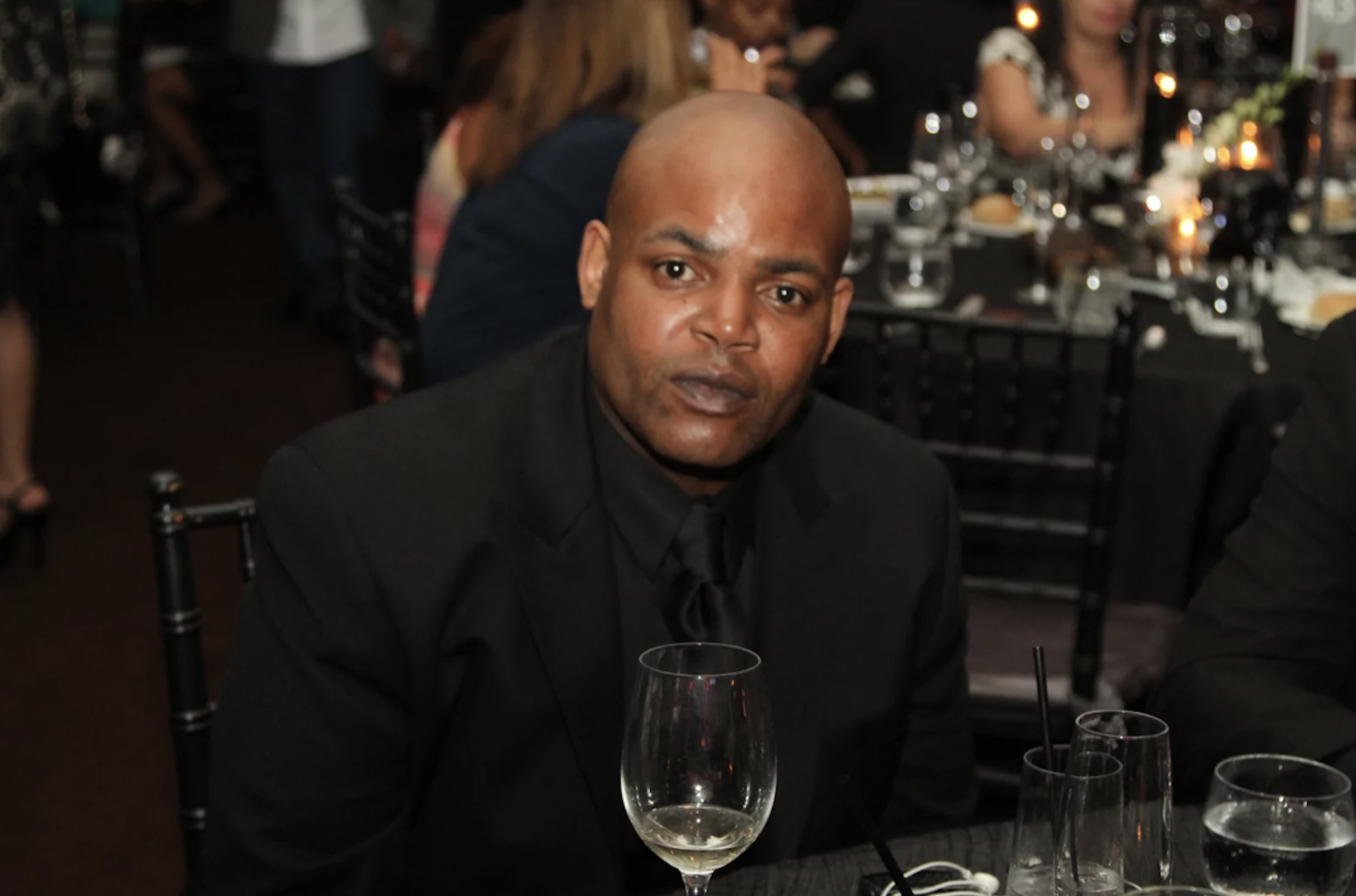 A bald Black man in a suit sits at a dinner table.