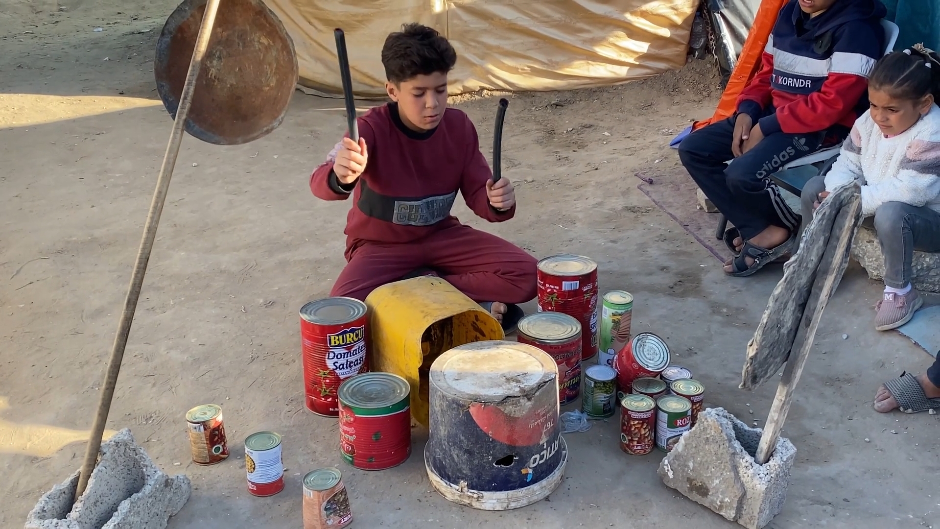 boy plays drums on an assortment of upturned buckets, others watch