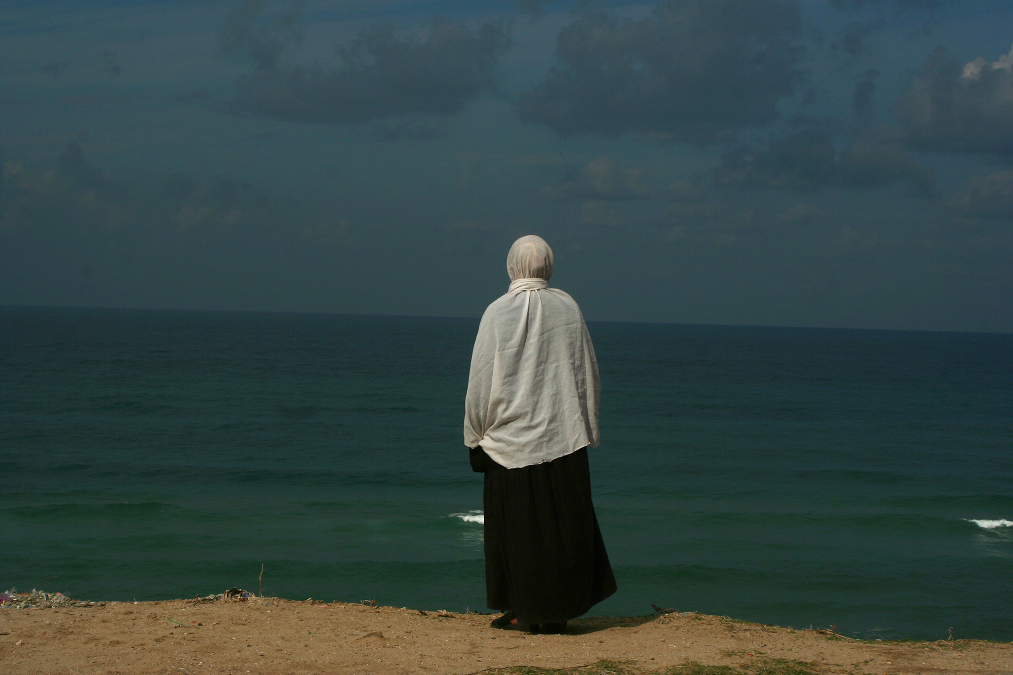 A photo of a woman in a hijab at a beach with her back turned, staring out onto the sea. 