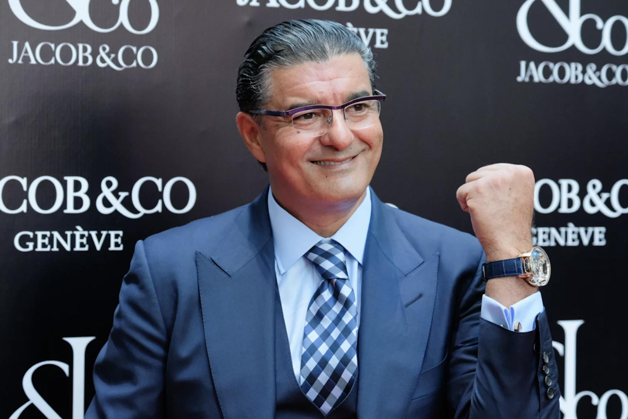 A middle aged white man in a blue suit holds one wrist up to show his watch.