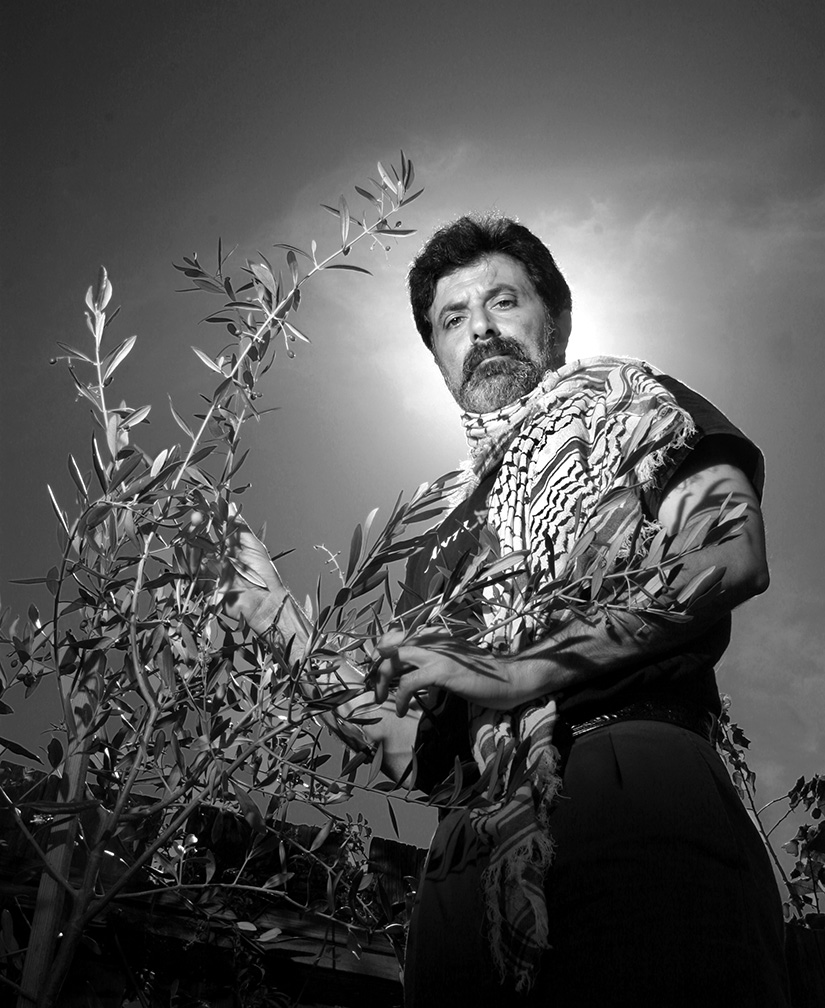 A black-andw-white self-portrait where the artist holds an olive branch while wearing a keffiyeh. 