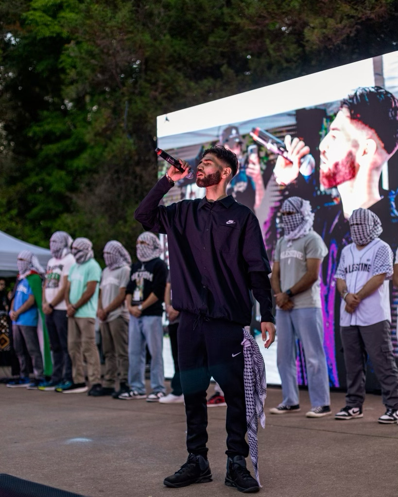 Sammy Shiblaq raps on stage with men behind him with their faces wrapped in keffiyehs.