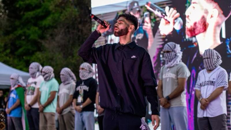 Sammy Shiblaq raps on stage with men behind him with their faces wrapped in keffiyehs.