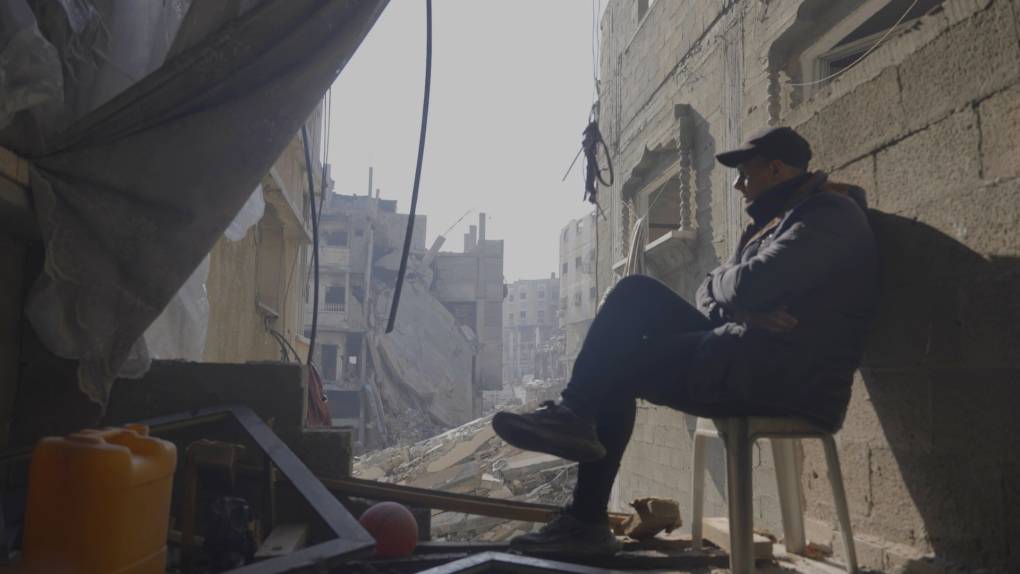 man sits on chair under lean-to, destroyed city of rubble in background