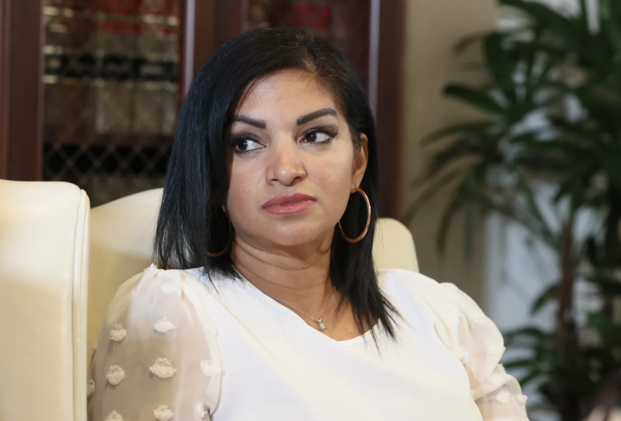 A light skinned Black woman sits on a white armchair wearing a white top.