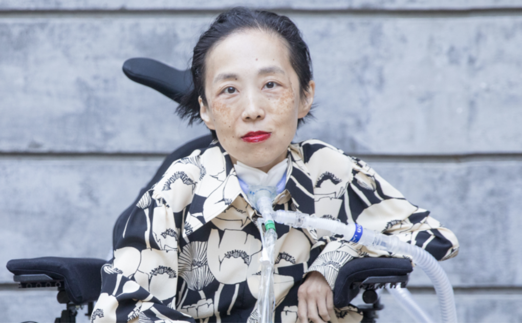 An Asian American disabled woman sits in a wheelchair with a tracheostomy with a tube attached to her ventilator. She is wearing a bold red lip color and a black and beige shirt with a pattern of poppies.