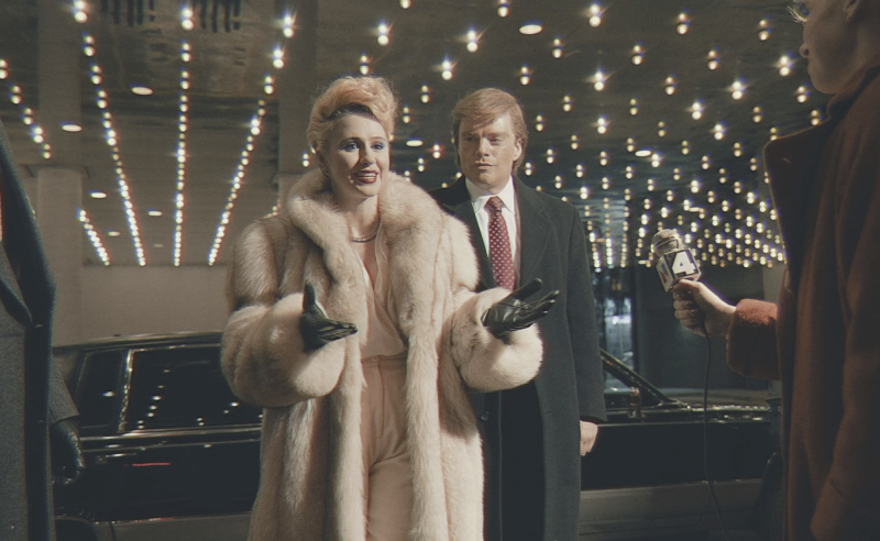 A woman in a fur coat and a man in a suit talk to a reporter outside a limo.