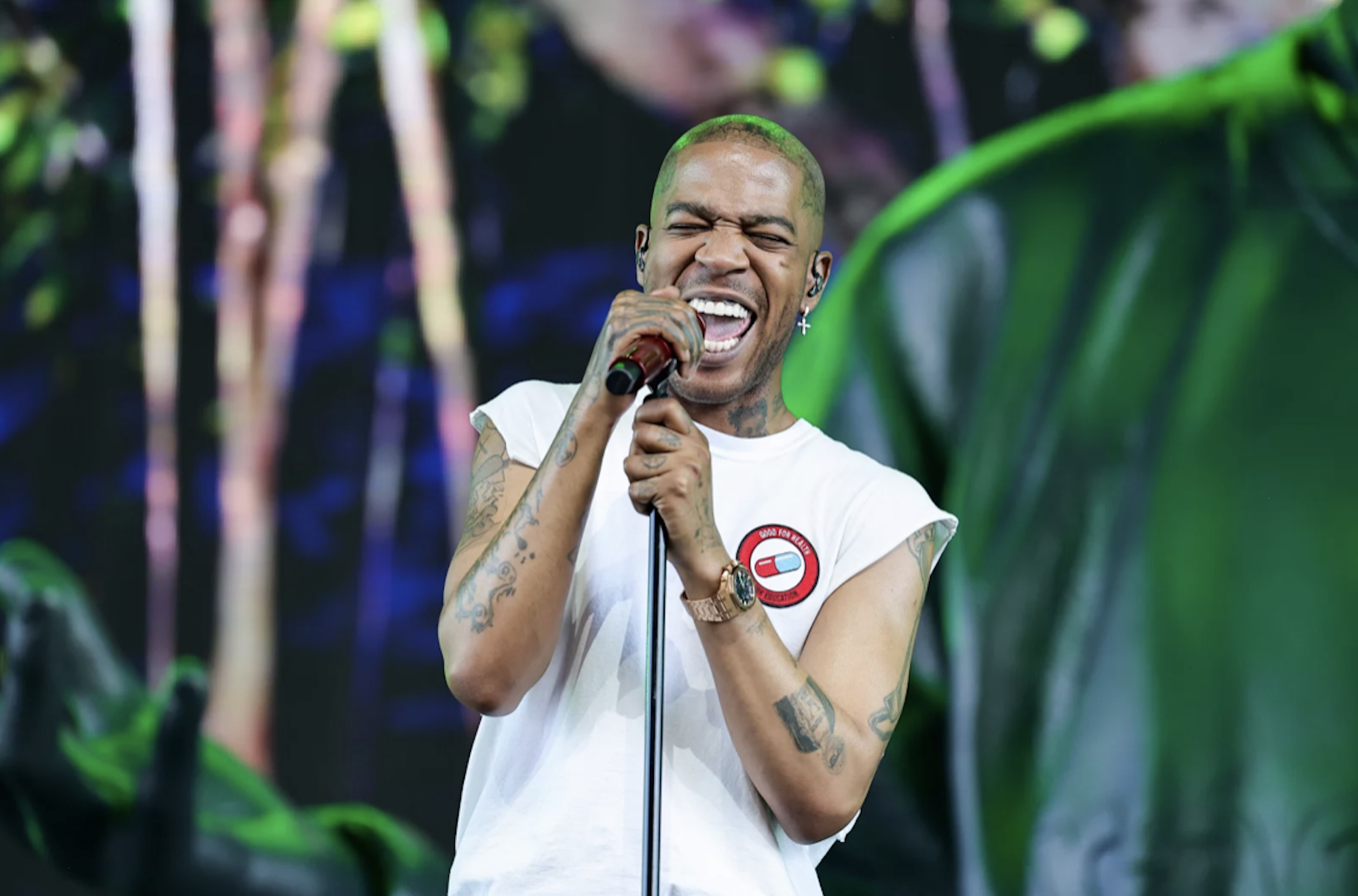 A Black man wearing a white t-shirt sings passionately into a microphone on stage.