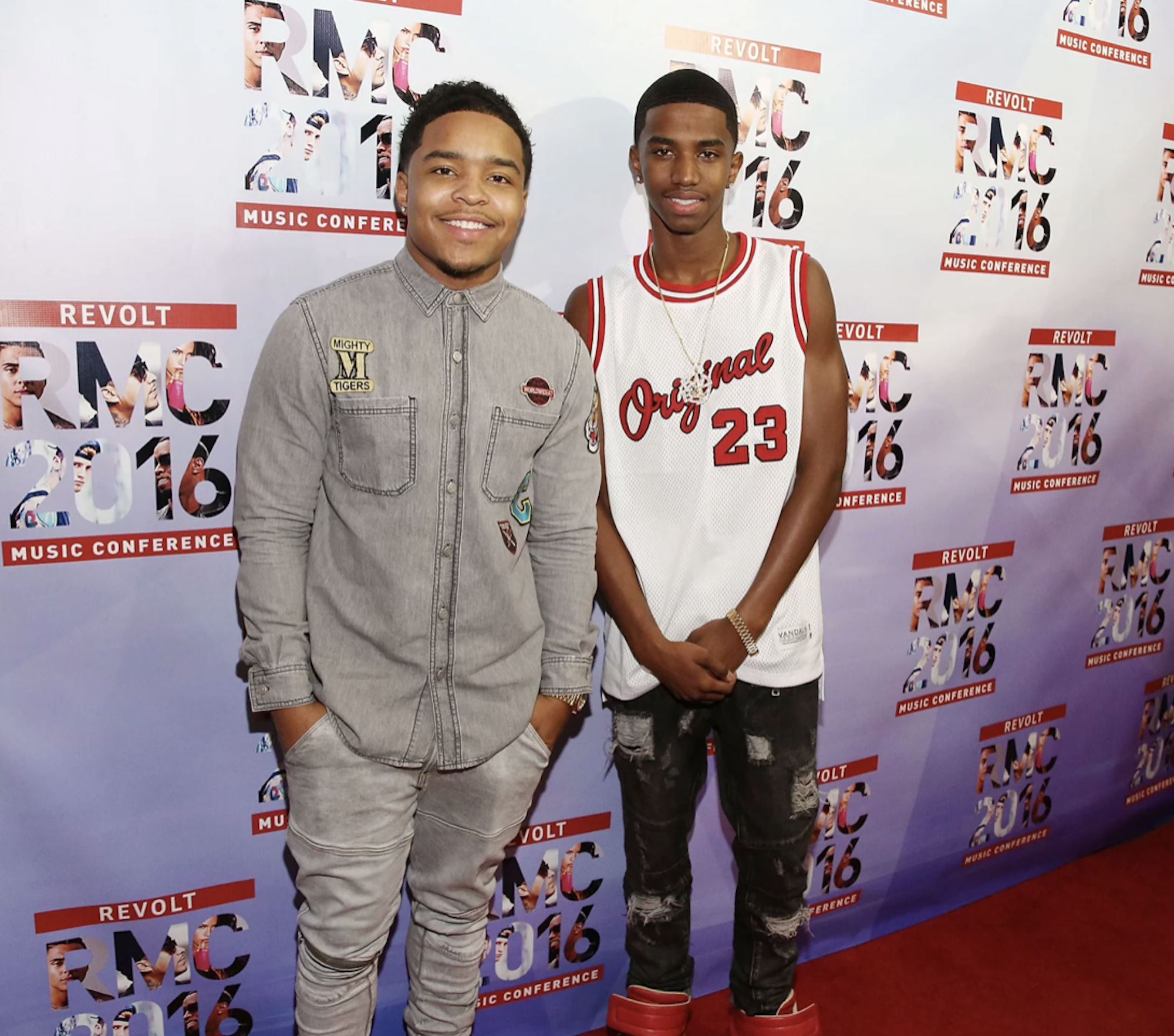 Two young, casually dressed Black men stand on a red carpet, smiling.