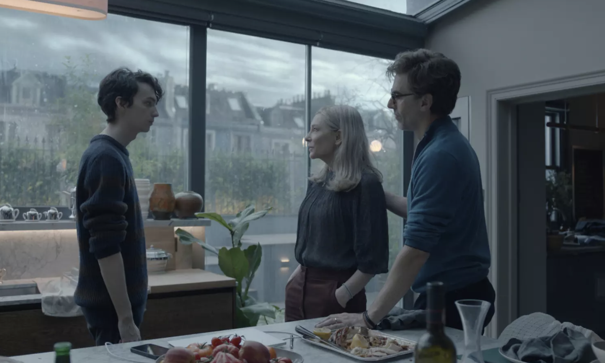 A man and woman stand in a kitchen with large windows talking to their adolescent son.