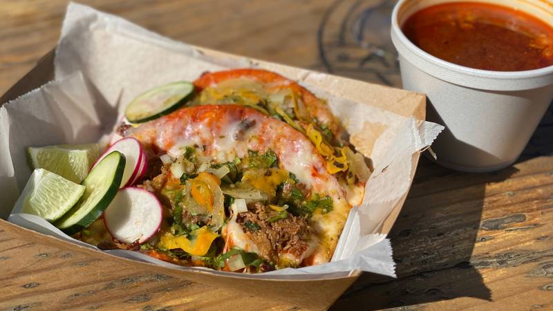 Two tacos topped with beef birria and melted cheese, with sliced cucumbers and radishes on the side.