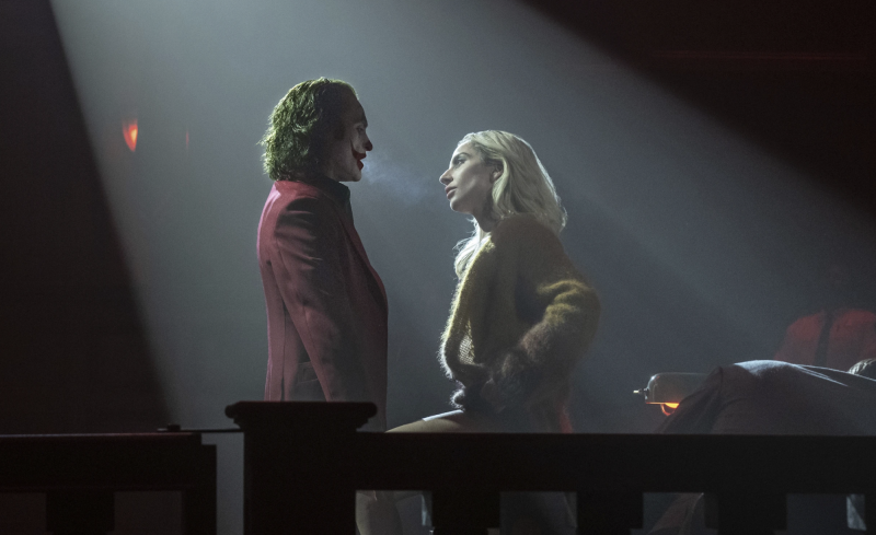 A man in red suit with green hair stands under a spotlight talking to a blond woman in a large empty dark room.