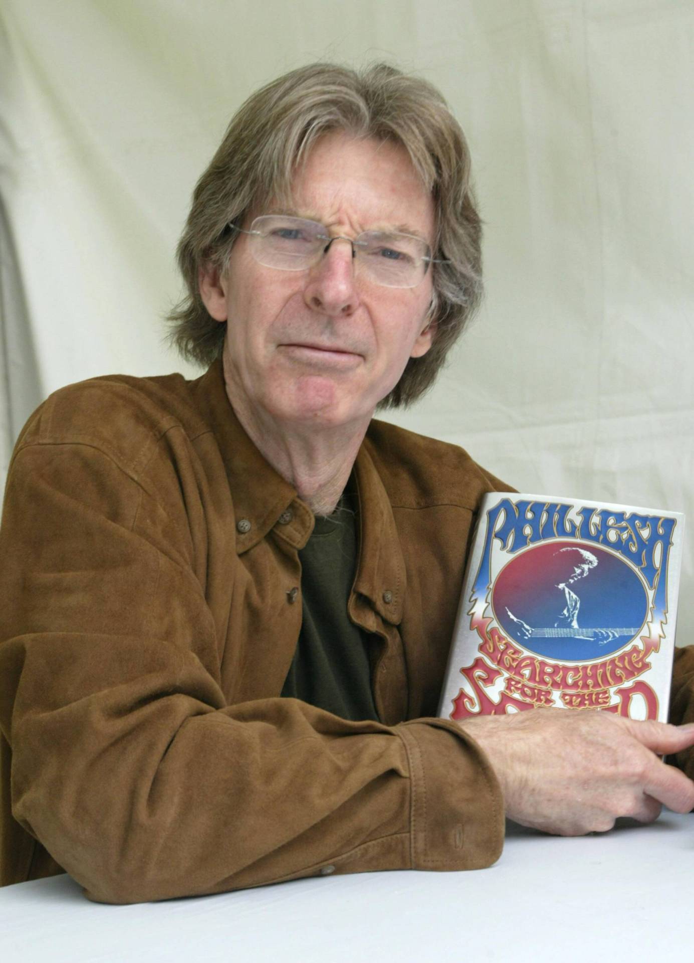 An older white man with grey hair wearing a brown suede jacket holding up a book with the Grateful Dead logo on it.