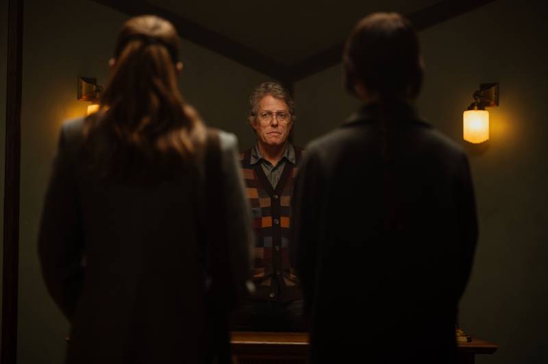 A grey-haired man wearing a shirt and cardigan stands before and between two female figures who are facing him.