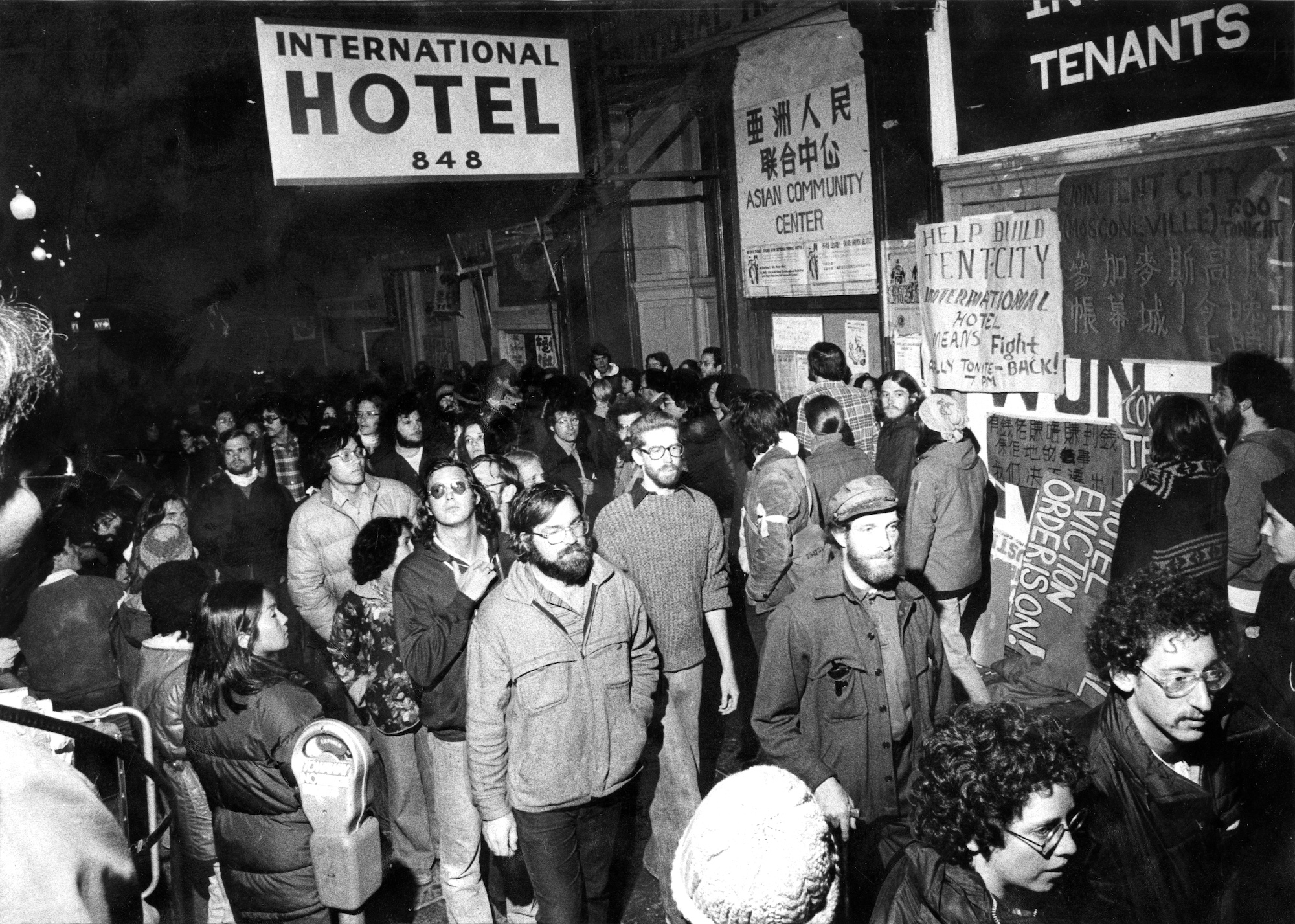 Men and women in 1970s clothing cluster together outside a building with a sign that says 'International Hotel' on it. 