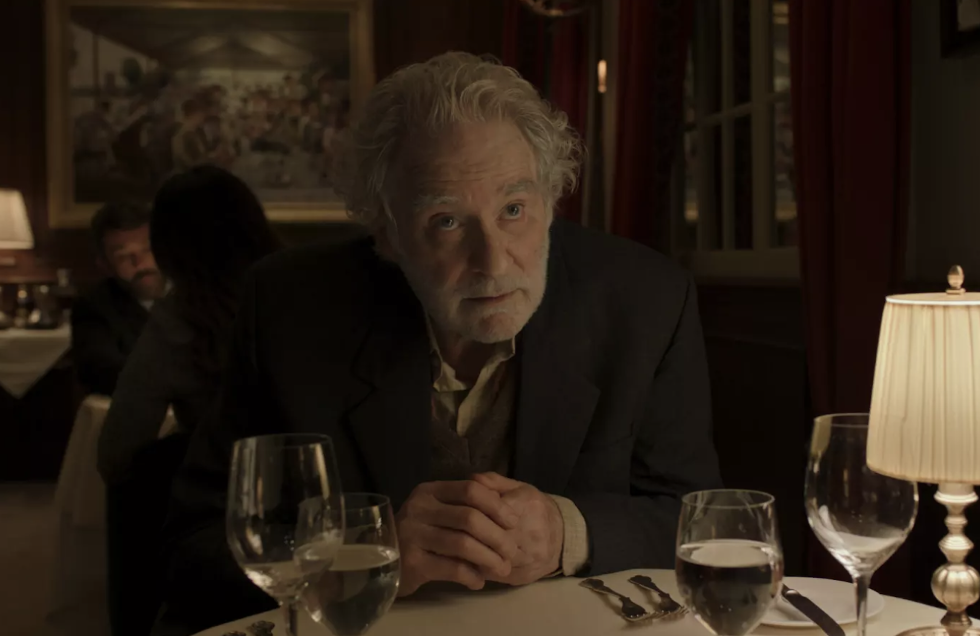 A man with white hair leans across a table in a dark bar or restaurant.