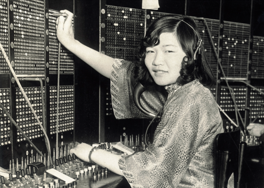 A young Asian woman operates a switchboard, while wearing a silk gown and telephone headset.