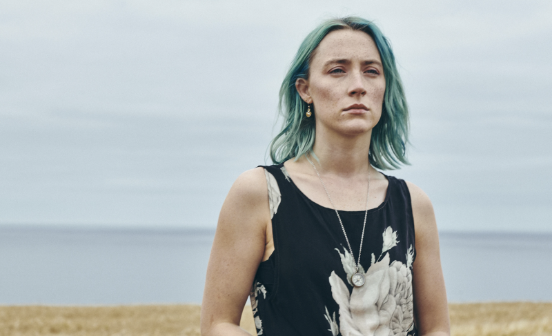 A gaunt white woman with blue hair stands on a hill, the sea visible behind her.