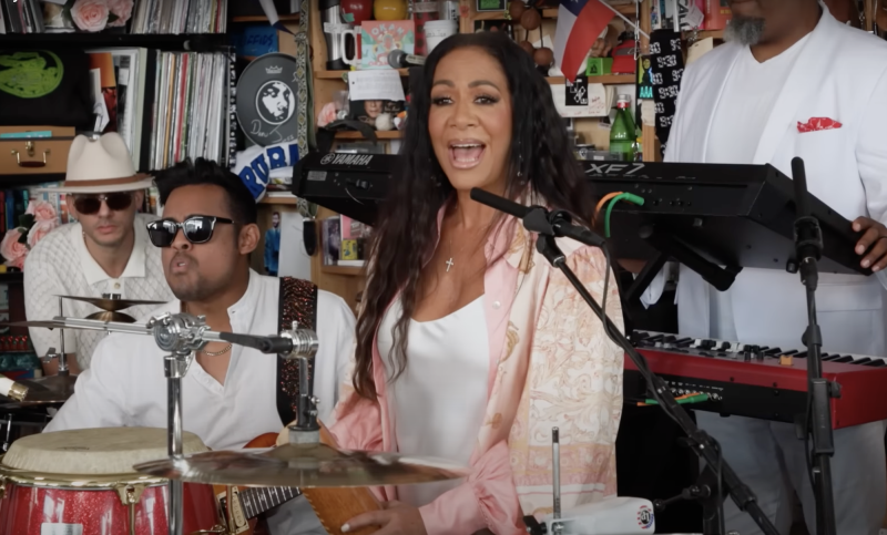 A Black woman with long flowing hair sings into a microphone, surrounded by musicians.