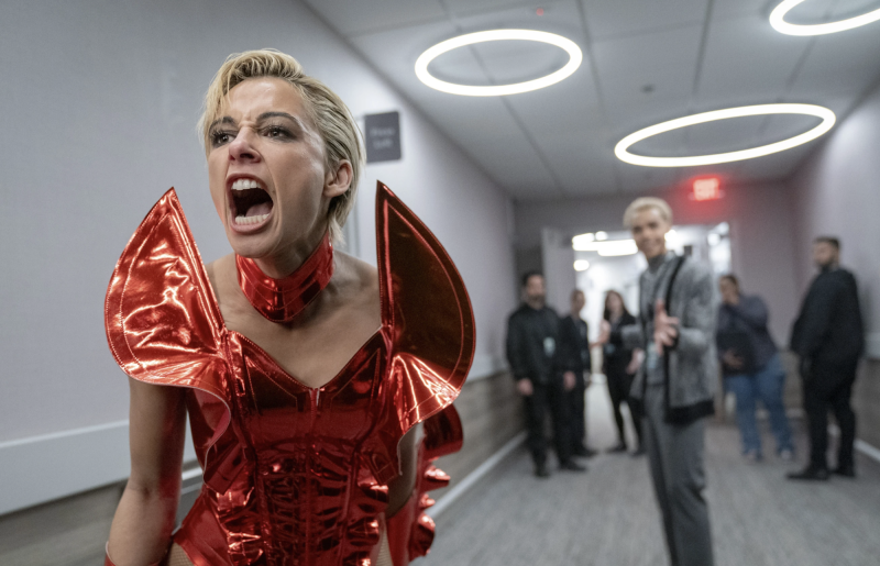 A woman in a striking red stage costume screams into a hallway, as onlookers stare at her from a distance.