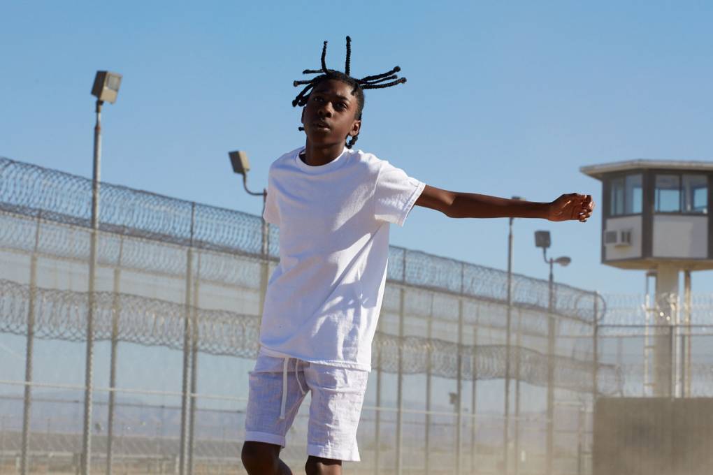 A scene from the film 'Songs From The Hole,' depicting a young African-American boy in a white shirt and white shoes dancing in a barbwire-lined prison yard.