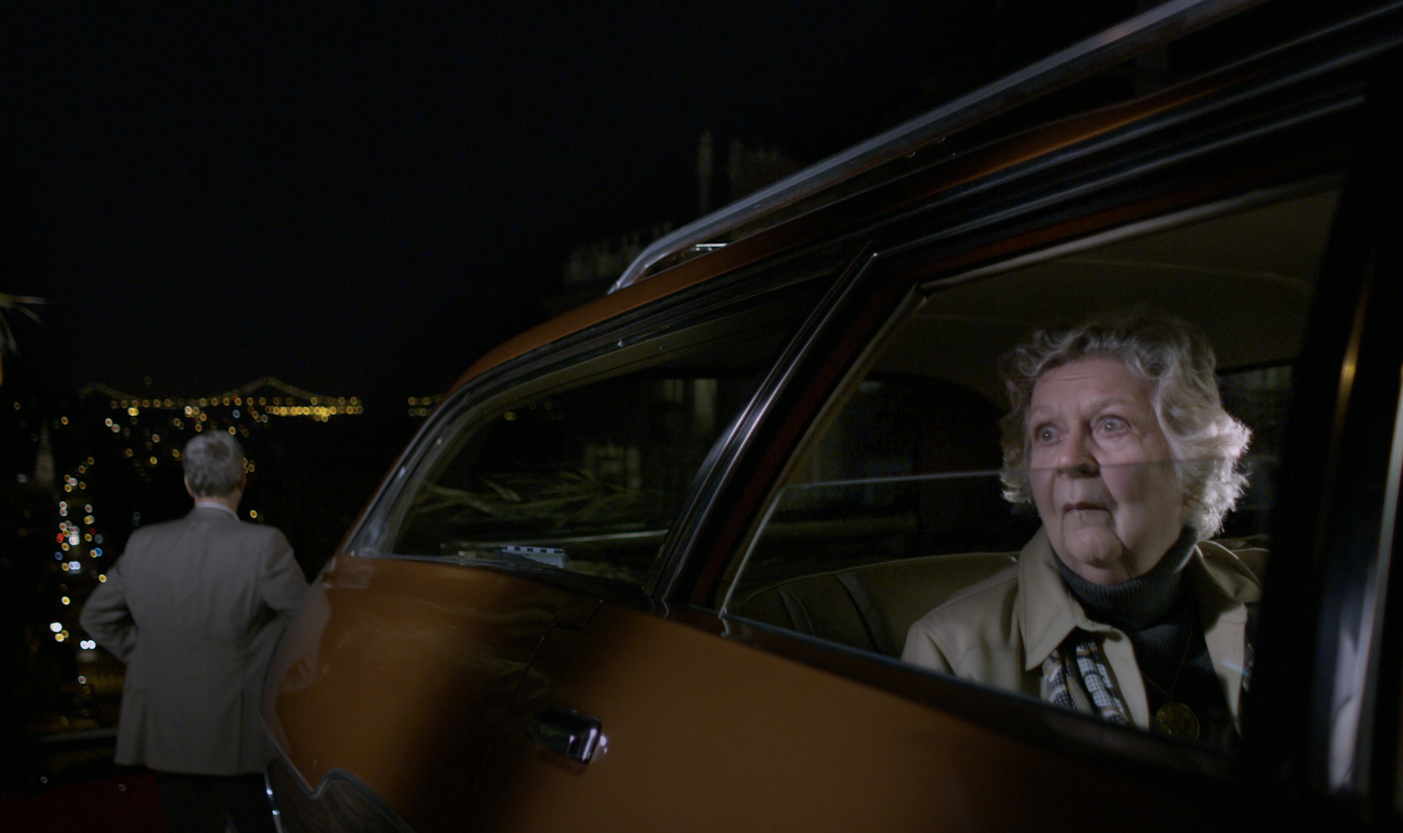 An elderly woman sits in the rear seat of the back seat of a Plymouth car, looking stunned. Behind the car, a man in a suit looks over a city skyline at night.