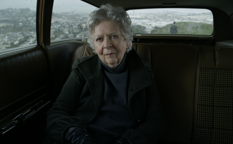 A senior woman, dressed all in black, sits in the back of a car on a hillside overlooking San Francisco.