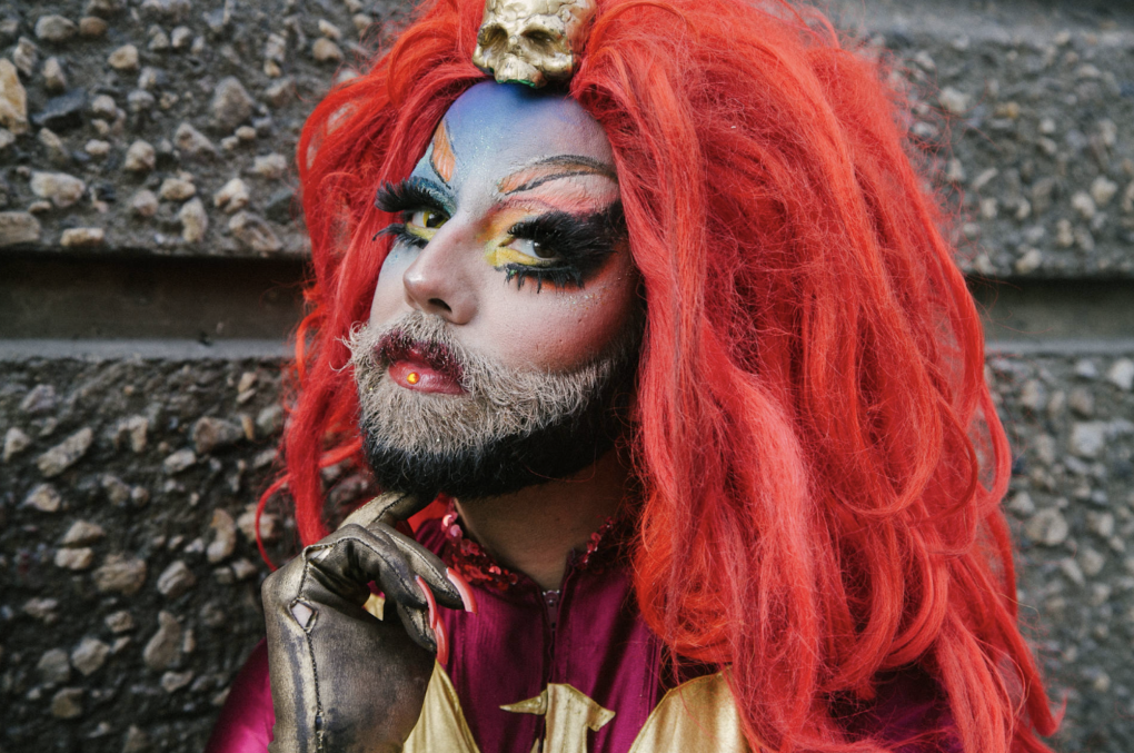 A bearded man in flamboyant makeup, large red wig and gloves with attached long fingernails.