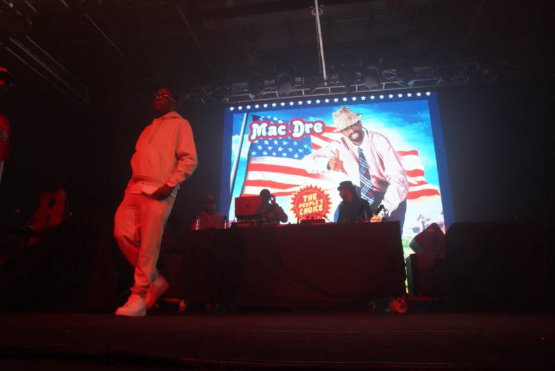 The image of Mac Dre's 'Ronald Dregan: Dreganomics' album cover is shown in the background, as veteran Vallejo MC B-Legit performs at History of The Bay Day in San Francisco. 