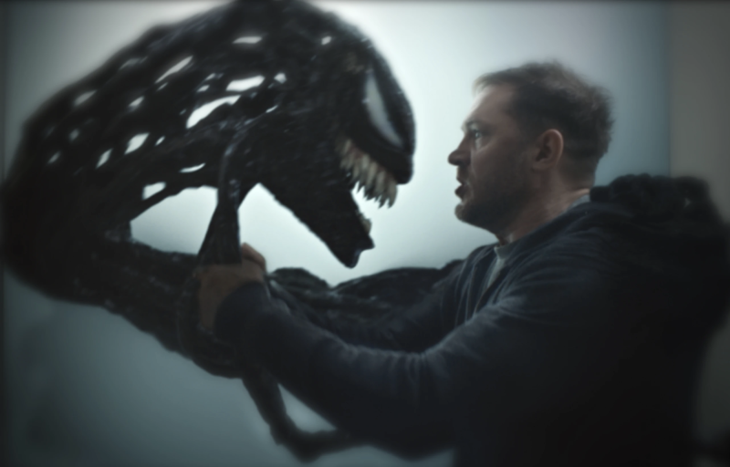 A white man in a black suit holds up and stares at a monstrous mask.