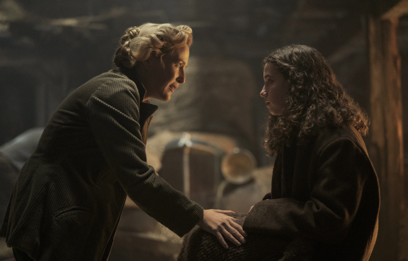 An adolescent girl sits inside a barn wearing an oversized coat. An older woman talks warmly to her.