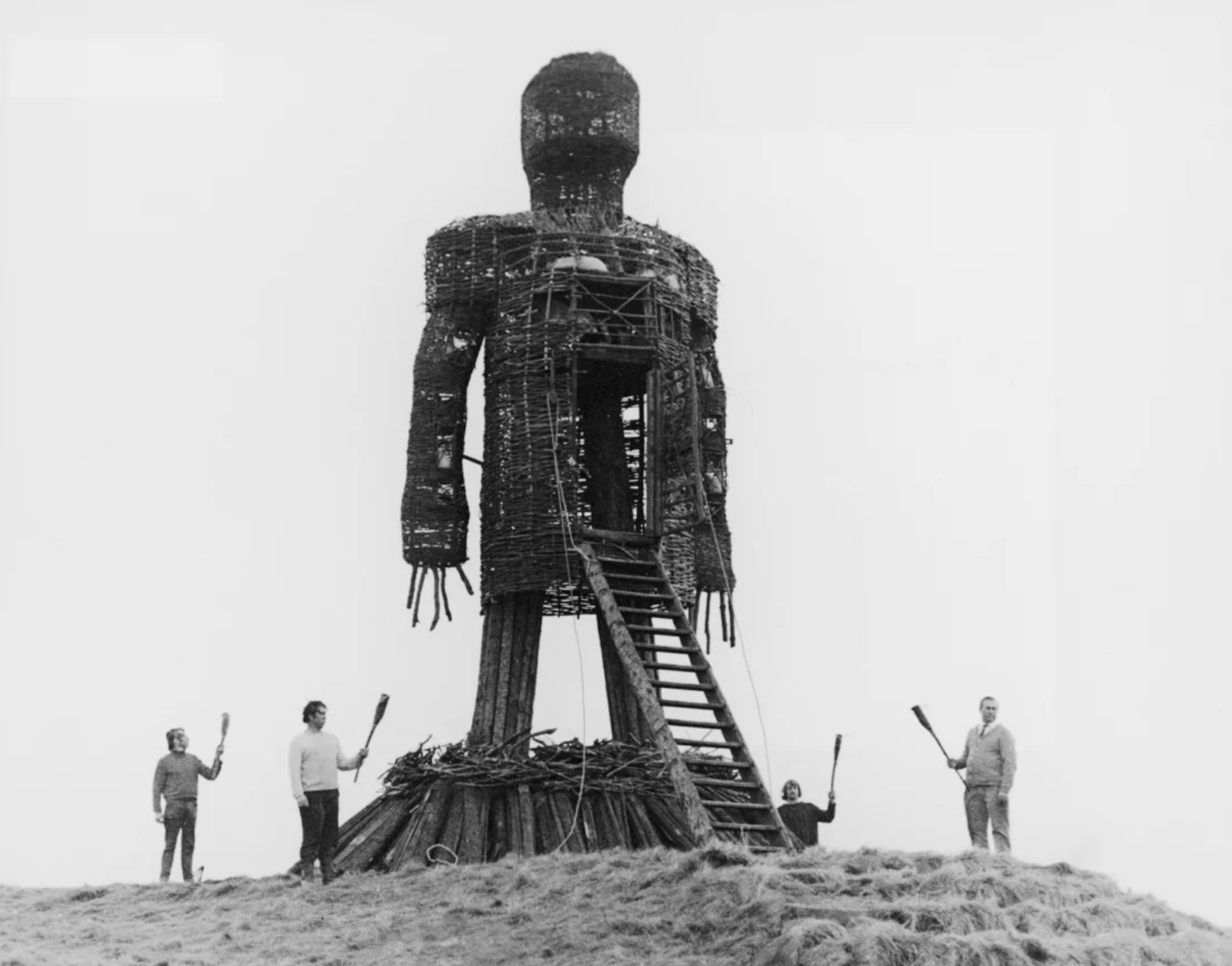 A huge wicker structure built in the shape of a man, stands on a hillside with a ladder propped up against its center. On the ground a circle of people with torches stand.