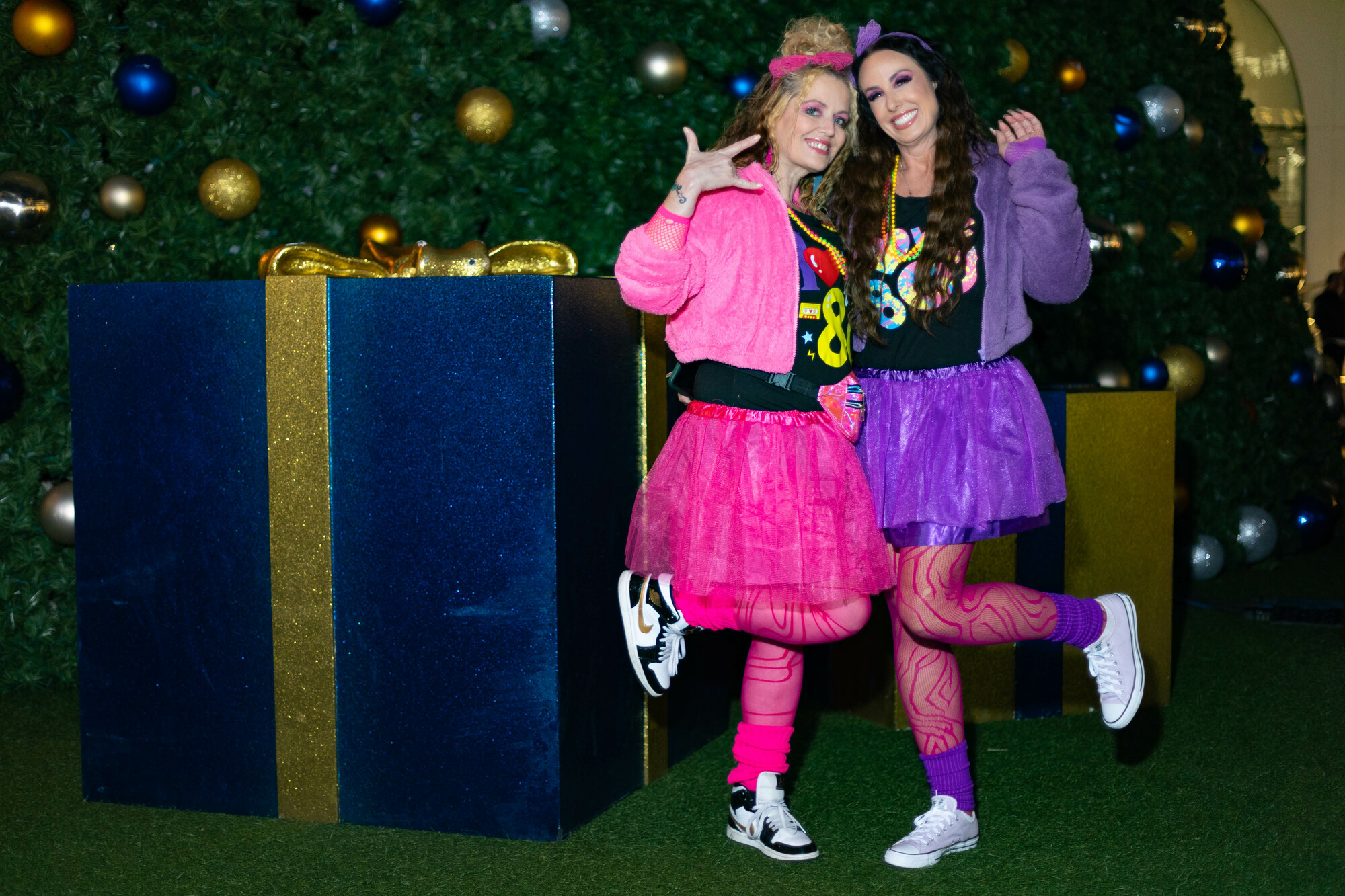 Two women in neon pink and purple outfits reminiscent of the 1980s pose together in front of a Christmas tree.