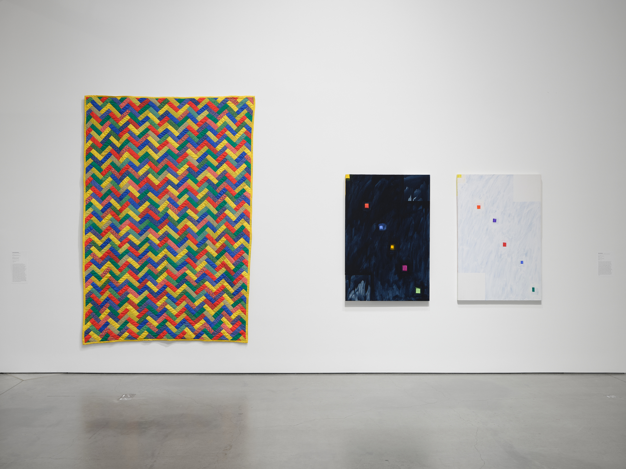 Large red, green, yellow and blue quilt with zig-zag pattern next to two paintings, one black and one white with a few colorful small squares.