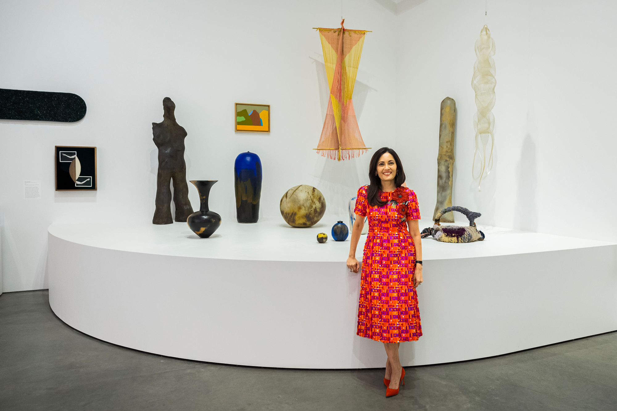 woman in bright red dress poses in front of high pedestal with ceramics and sculptures on it