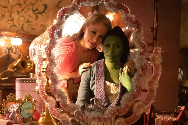 A white blond woman dressed in pink rests her hand on the shoulder of a green woman dressed in black sitting at a vanity mirror.
