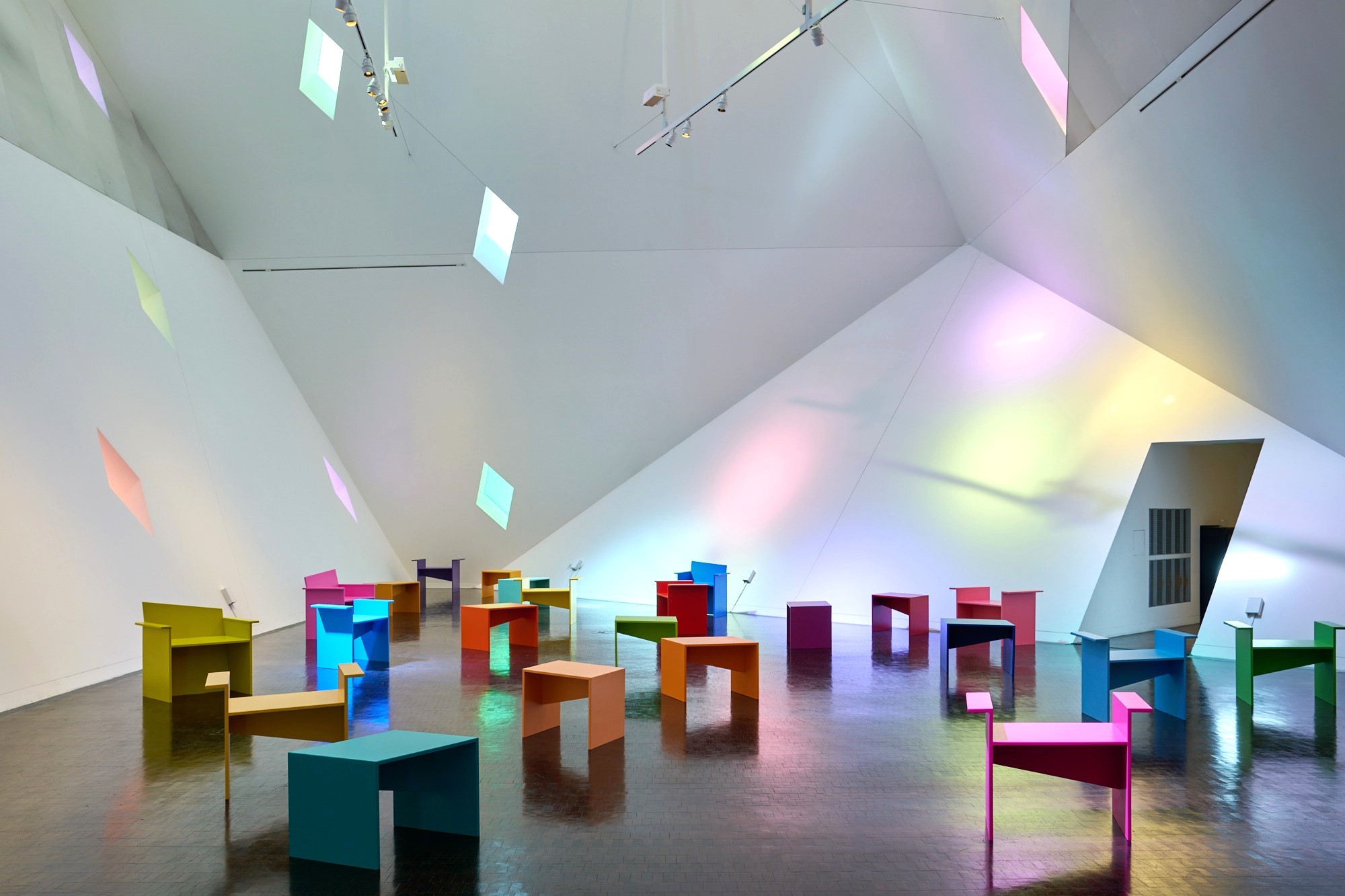 colorful chairs and benches in an angular space with colored light cast against walls