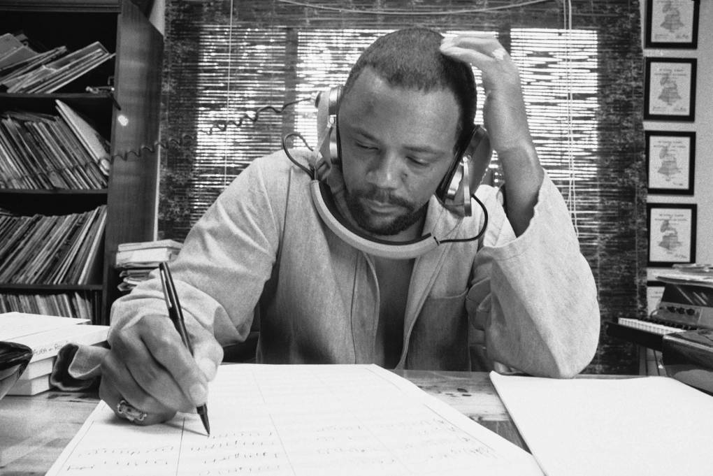 A black man wearing a sweater and headphones notates music on a music staff at his desk, with vinyl LPs on a shelf in the background