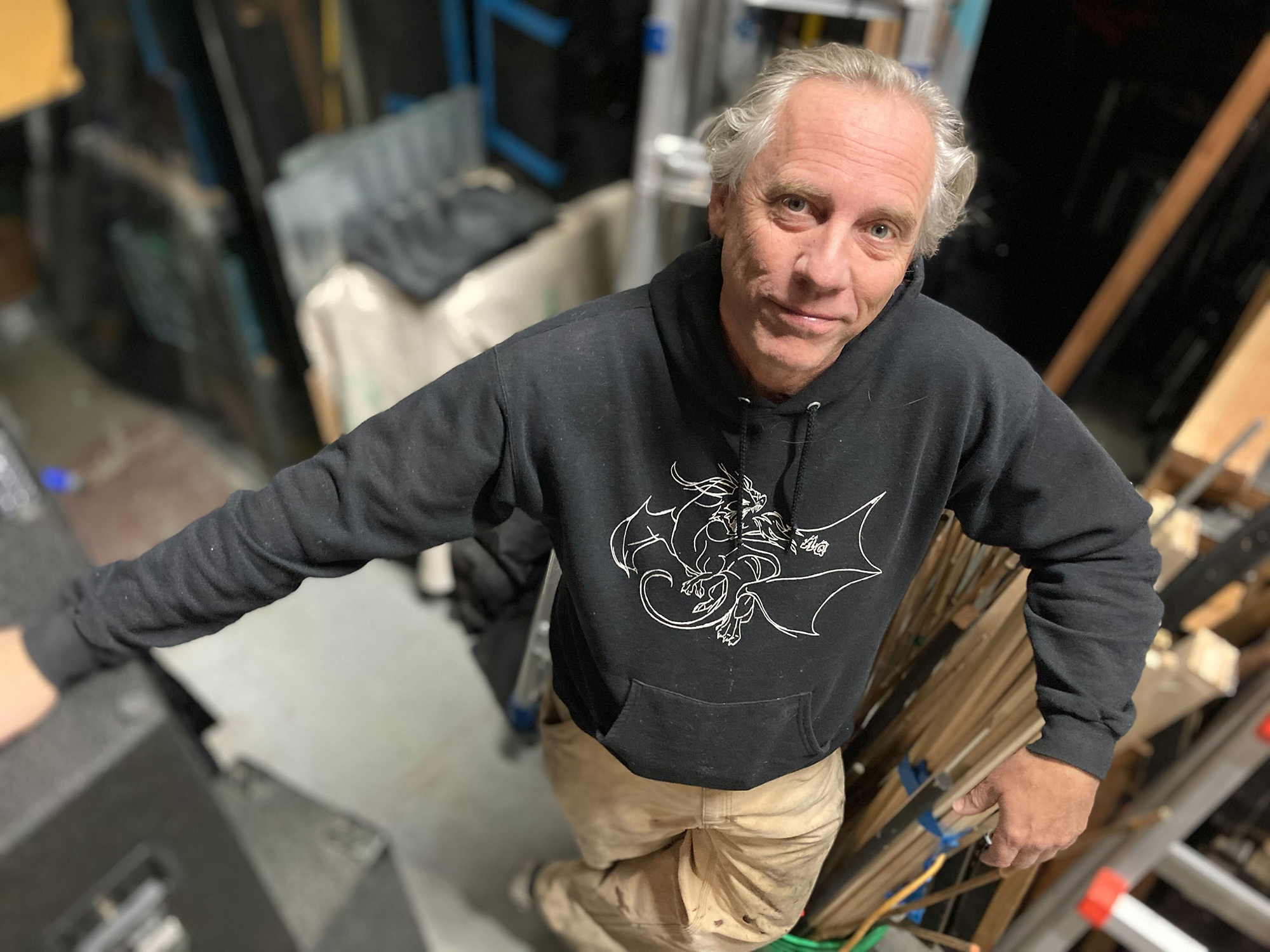 A man in a black hoodie and tan pants stands among a cluttered studio space, looking up at the camera.