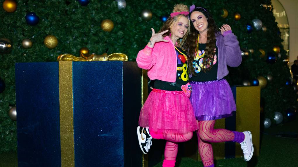 Two women in neon pink and purple outfits reminiscent of the 1980s pose together in front of a Christmas tree.
