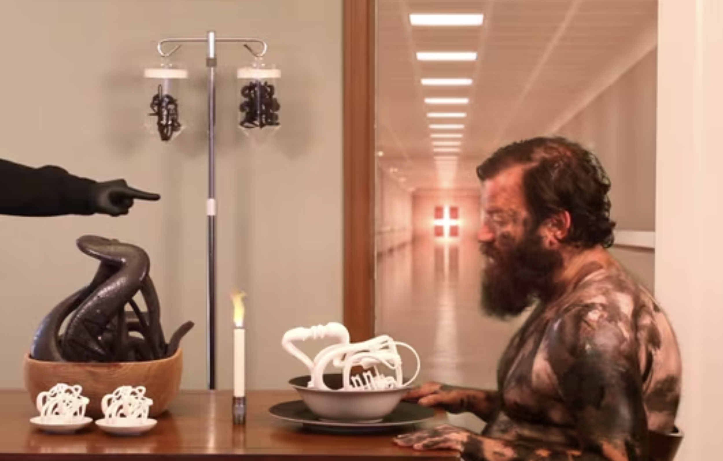 man with beard covered in dark substance sits at table with abstract sculptures in dishes, pointing hand emerges from left