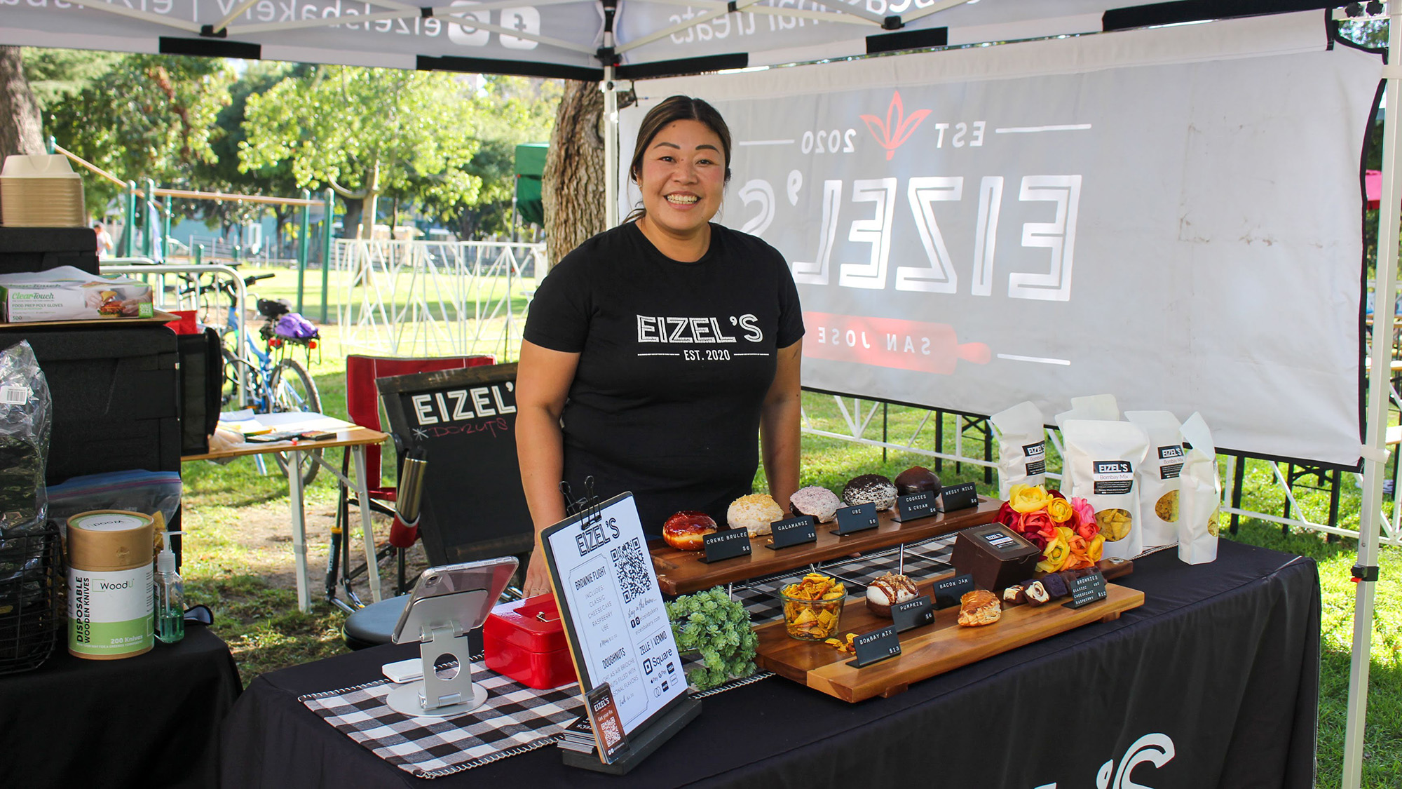 A baker poses at her farmers market display of doughnuts and other baked treats.