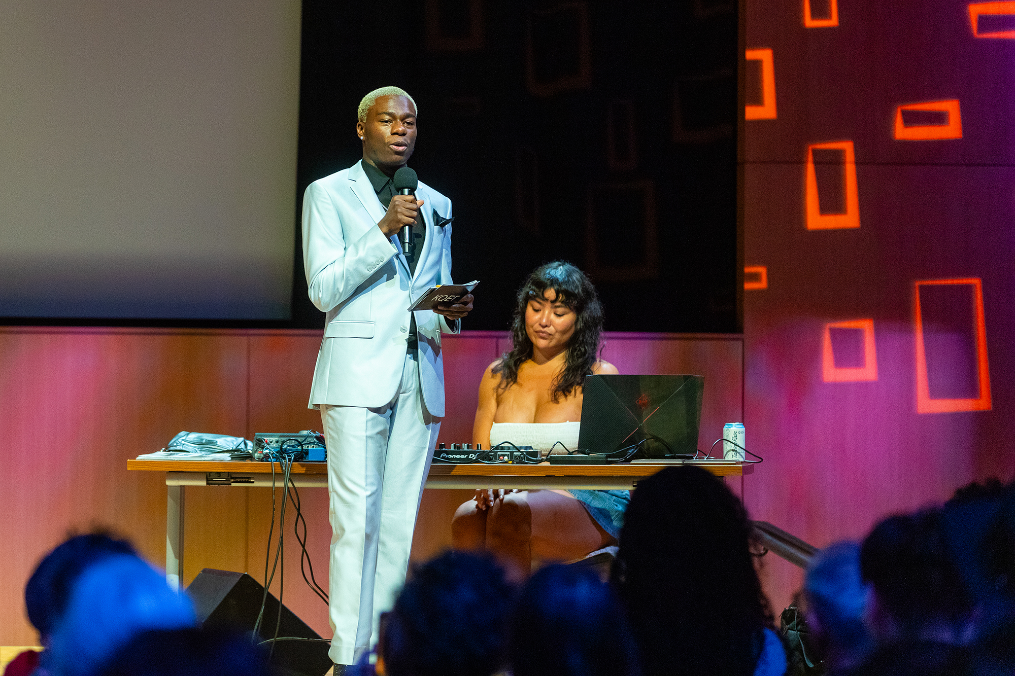 One person wearing a suit and holding a microphone stands on an indoor stage in front of a crowd. There is another person to the side sitting in front of a DJ table.