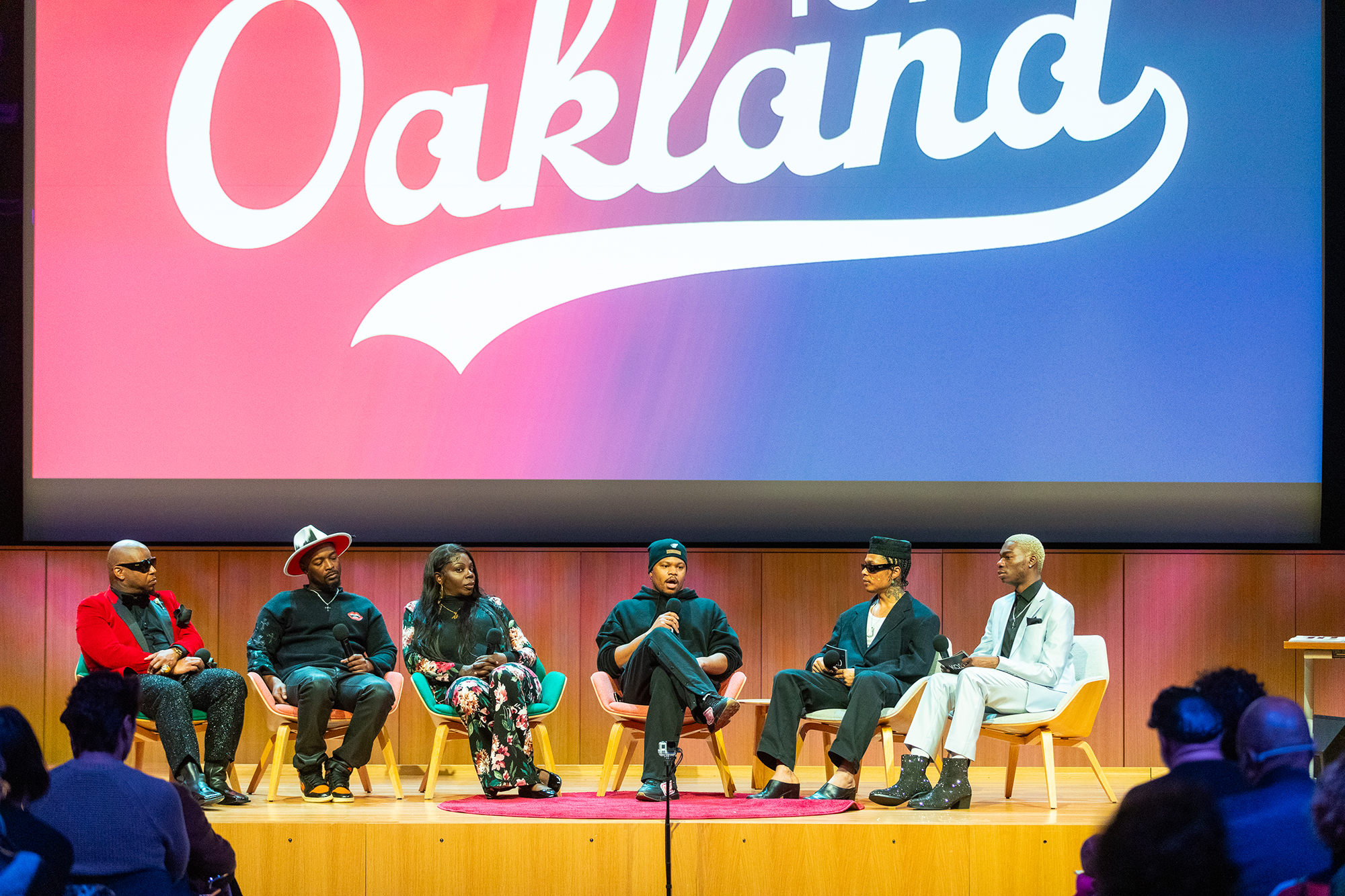 Six people sit on chairs on an indoor stage with a large projection behind them that reads, "Oakland to All."