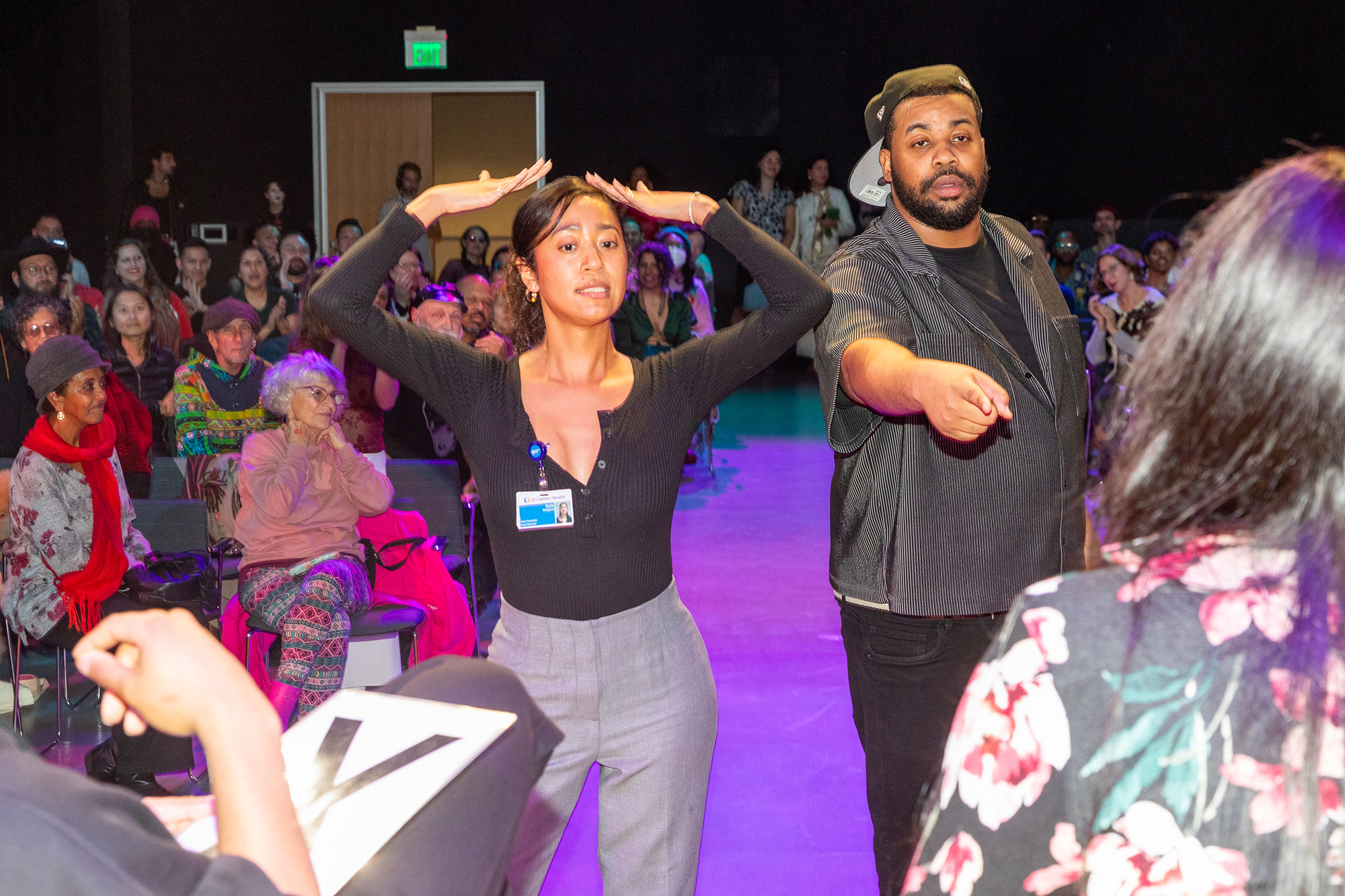 Two people perform in front of a group of judges that are sitting down inside an indoor auditorium. A large crowd sits behind them.