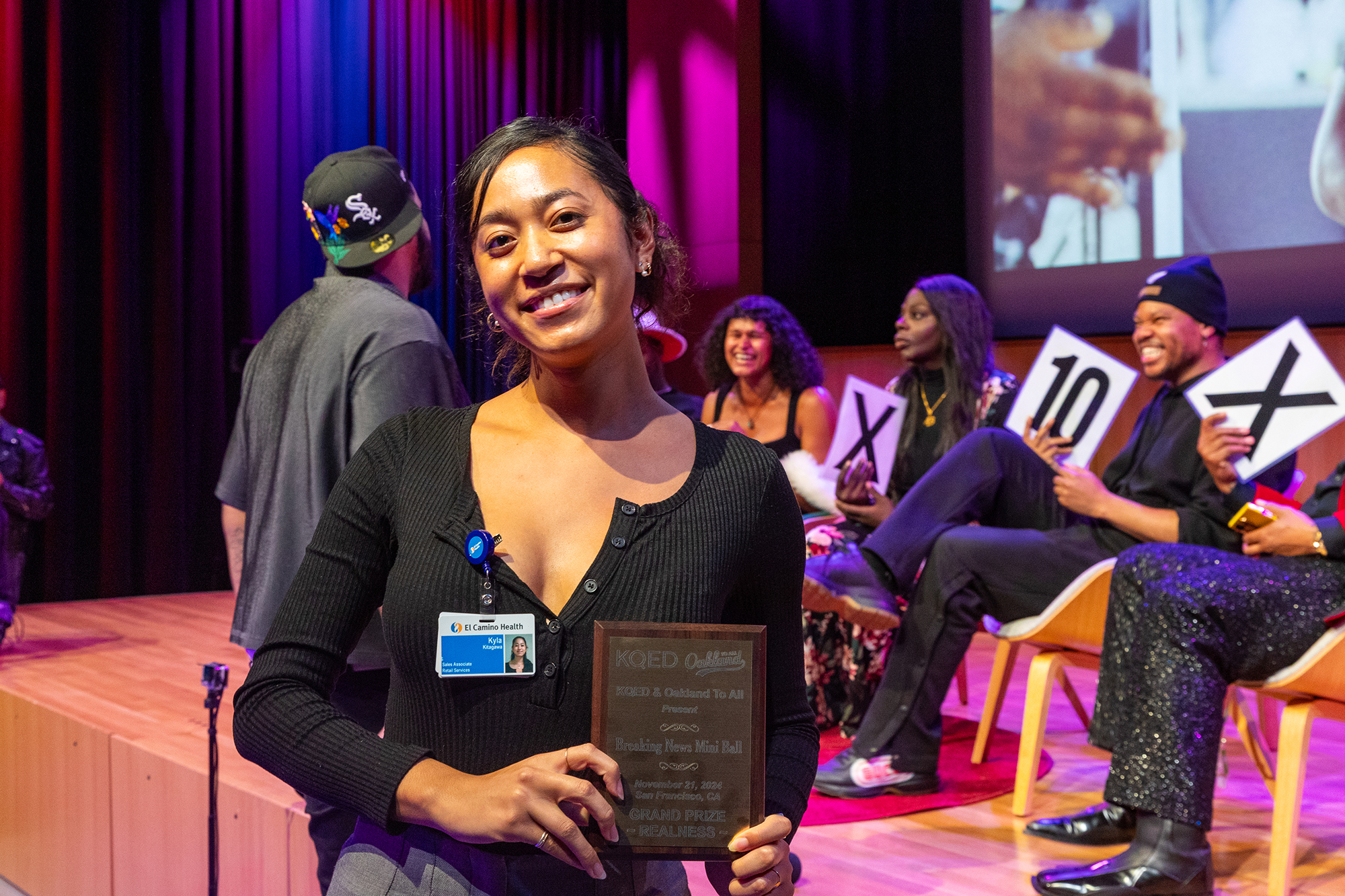 A person holds up a plaque in a crowded auditorium and smiles at the camera.